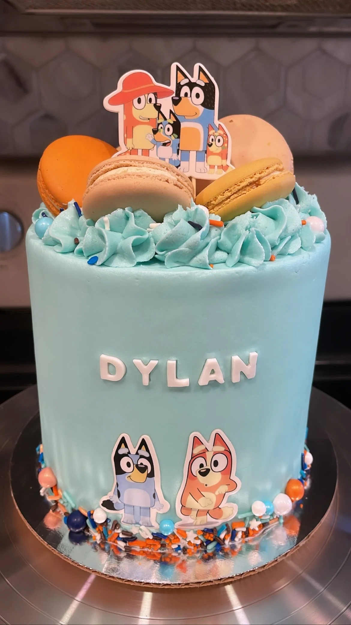 Bluey themed birthday cake in light blue with character images, macarons, and colorful sprinkle border